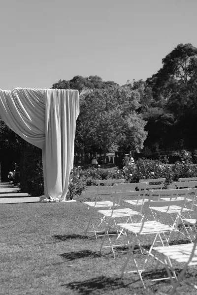 Outdoor ceremony setup with a draped ceremony arch and white folding chairs on a grass lawn framed by garden trees and rose bushes.