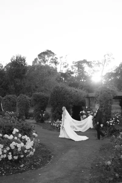 Couple portrait of a bride and groom walking along a garden path, her veil trailing as rose bushes frame a pergola under the open sky.
