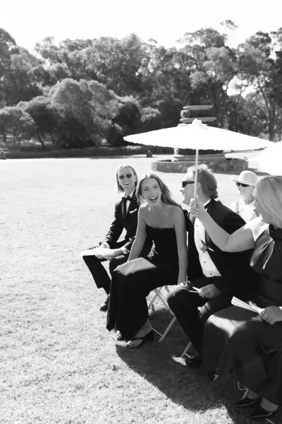 Ceremony guests gather as outdoor wedding guests laugh under parasols on a garden lawn near a stone fountain.