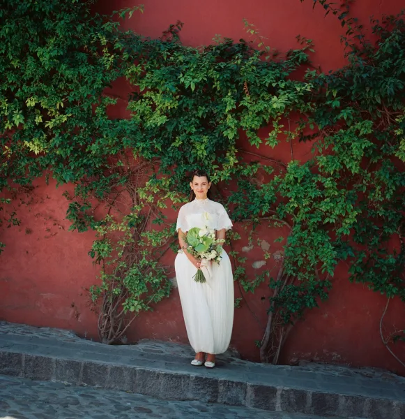 Bridal portrait of a bride with bouquet, wearing a lace top, framed against a red wall with climbing vines along a cobblestone path.