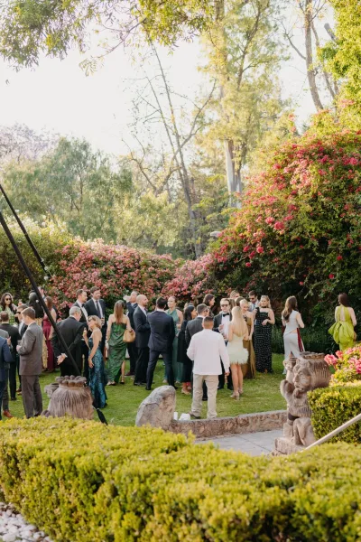 Wedding cocktail hour with wedding guests mingling on a garden lawn, champagne flutes in hand, framed by flowering hedges and stone sculptures.