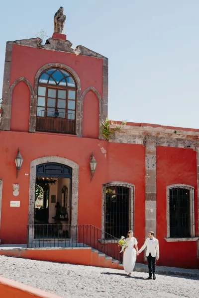 Couple portrait of a bride and groom walking hand in hand as she holds a greenery bridal bouquet by a red stucco building with arched windows.