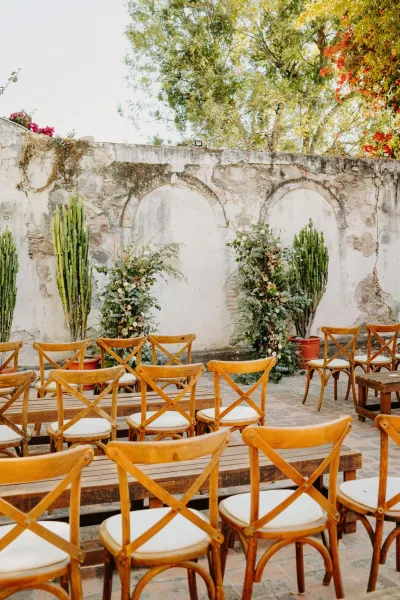 Ceremony setup featuring outdoor ceremony seating with cross-back chairs facing a stone wall and bougainvillea along a brick patio garden.