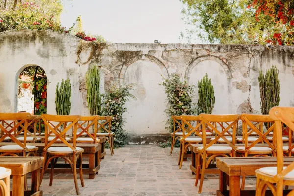Ceremony setup showcasing courtyard ceremony decor, wooden chairs facing a stucco wall with arched niches and a terracotta tile floor.