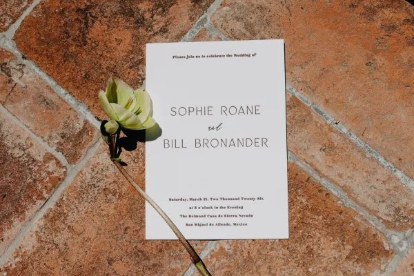 Wedding invitation in a minimalist wedding invitation layout on terracotta tile with grout lines, accented by a single flower.