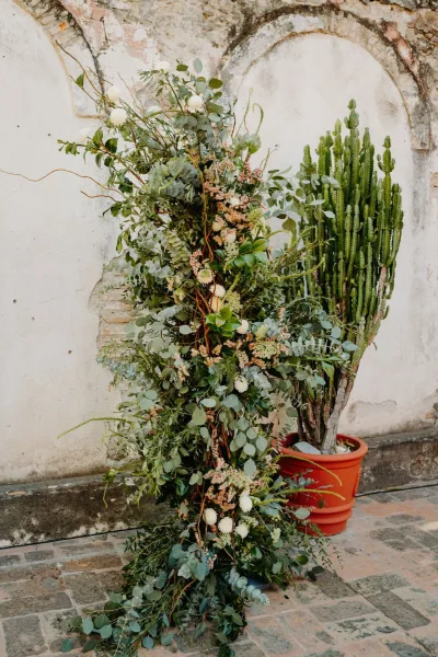 Ceremony floral pillar with eucalyptus rises beside a stone arch against a plaster wall, set on brick pavers.