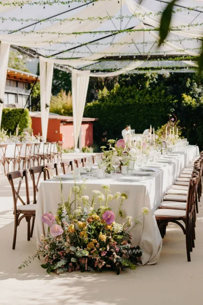 Reception tablescape for an outdoor reception table, taper candles glowing down the center beneath a tent frame beside garden hedges.