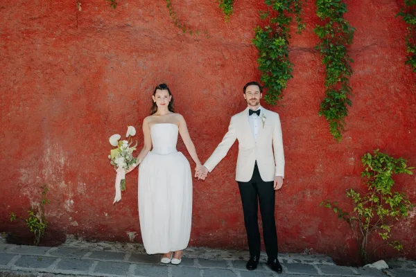 Couple portrait of bride and groom holding hands, the bride cradling calla lilies against a red wall with green vines and stone pavement.