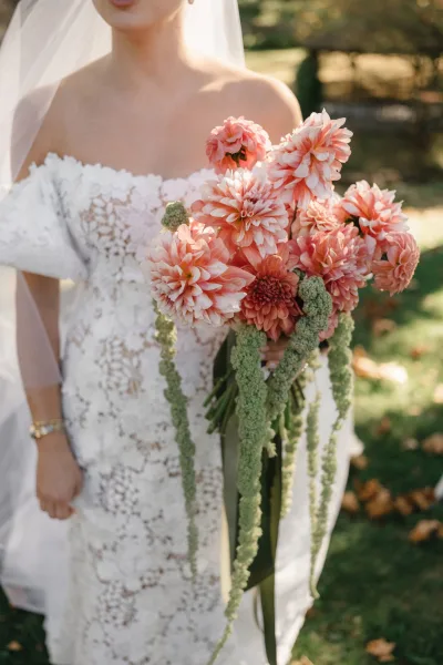 Bridal bouquet with dahlia wedding bouquet blooms and cascading greenery held by bride in strapless lace dress on a sunlit lawn with trees