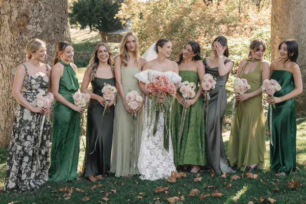 Bridesmaid group photo with bride and bridesmaids in green dresses holding white and blush bouquets on a sunlit garden lawn among trees