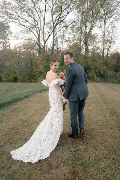 Couple portrait of bride and groom holding hands, walking away and looking back, bride’s lace dress train and pink bouquet in a grassy field