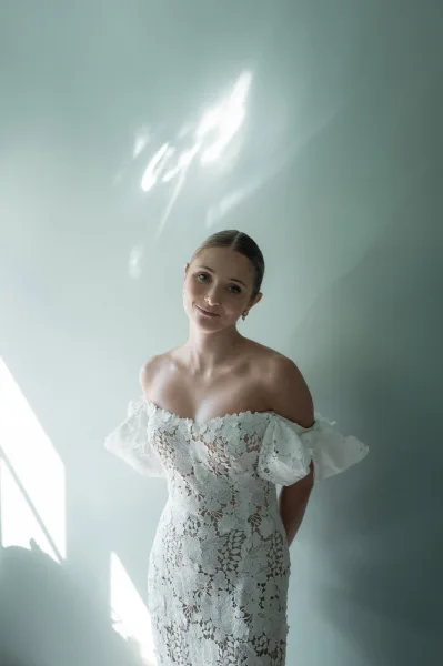 Bridal portrait of a bride in an off the shoulder wedding dress with lace details and drop earrings, lit by window light on a plain wall
