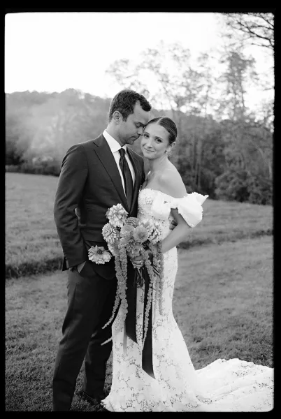 Couple portrait in a black and white wedding portrait as groom kisses bride’s forehead; she holds a cascading bouquet with long ribbons in a grassy field