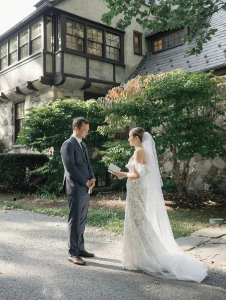 First look moment as bride in lace gown and veil reads vows from a book to groom in suit on a driveway by a stone house