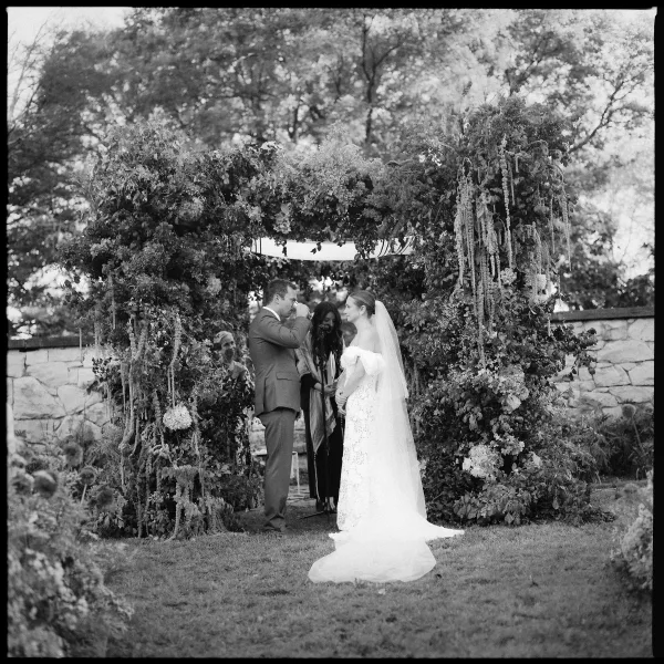 Wedding vows at an outdoor wedding ceremony as bride in veil and groom in suit face each other under a floral arch before a stone wall