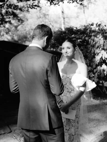 Wedding vows as bride in lace dress reads from a vow book to groom in suit, veil accent, in a dappled sunlight garden ceremony