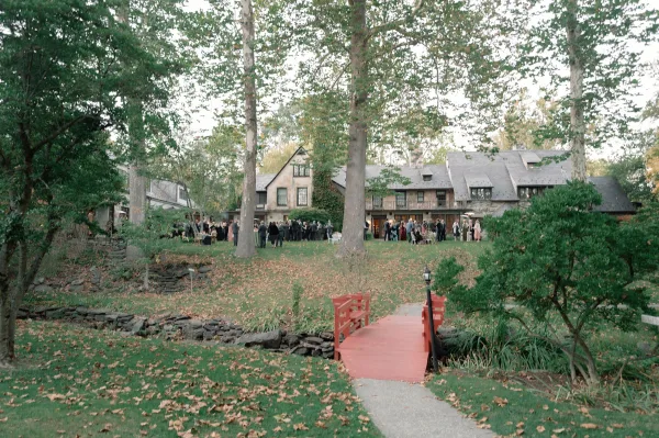 Wedding cocktail hour on an estate lawn with outdoor cocktail tables and heaters, near a creek and footbridge amid fallen leaves and tall trees