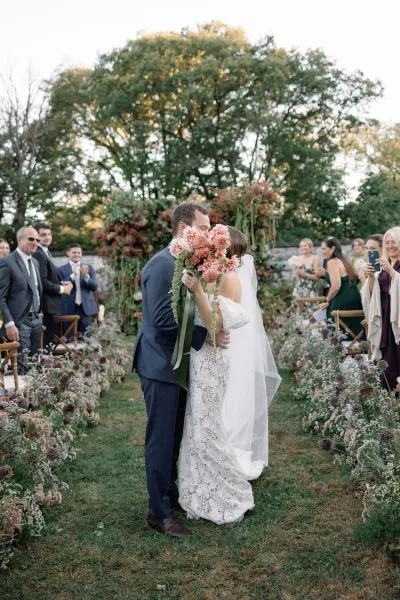Wedding kiss as bride and groom kiss behind a bridal bouquet with long ribbon, on a garden aisle with wooden chairs and guests