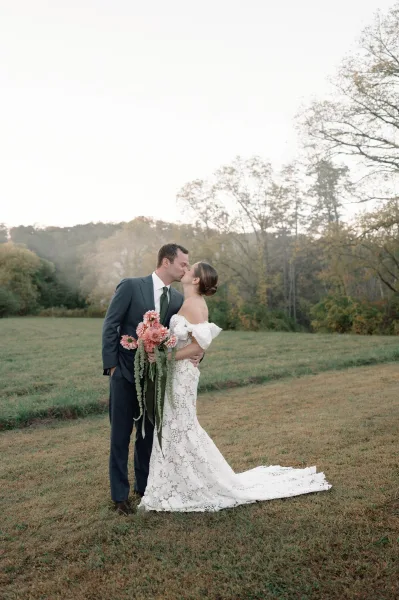 Wedding kiss portrait of bride and groom kissing as she holds a pink bouquet with trailing greenery, full length in a grassy field with hills