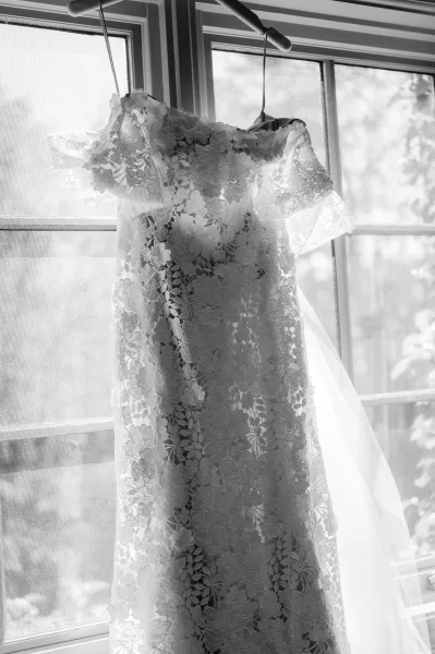 Wedding dress, lace wedding dress hanging on a hanger in a window with a tulle veil draped beside it, trees faintly visible outside
