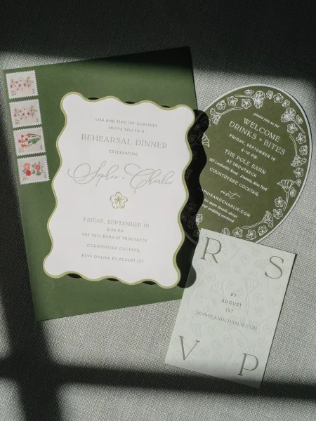 Wedding invitation suite with green wedding invitations, scalloped and round cards, RSVP, and floral postage stamps on gray fabric in sun shadows