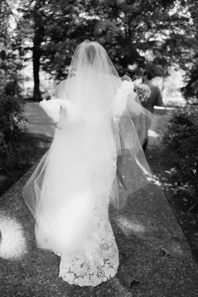 Wedding first look as bride walks to groom on a tree-lined garden path, holding a white bouquet behind her lace dress and long veil
