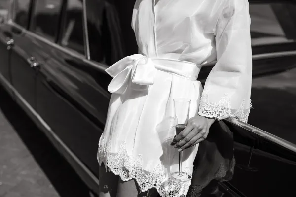 Bridal getting ready moment with a bride in robe holding a champagne flute, lace-trimmed white satin sash bow beside a black car door