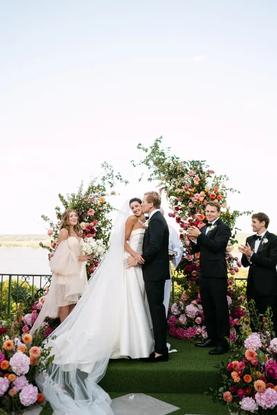 Ceremony kiss beneath a floral ceremony arch as bride in strapless dress and long veil kisses groom in tux on an outdoor terrace