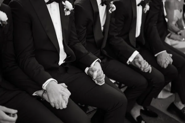 Groomsmen portrait with groomsmen in tuxedos sitting with hands clasped, black bow ties and boutonnieres, guests seated behind