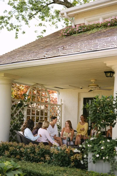 Bridal party getting ready as bridesmaids relax in robes with champagne flutes on a covered porch with trellis and greenery backdrop