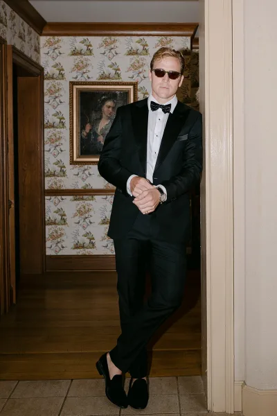 Groom portrait in a black tuxedo with bow tie and sunglasses, leaning in a doorway against floral wallpaper and framed painting indoors