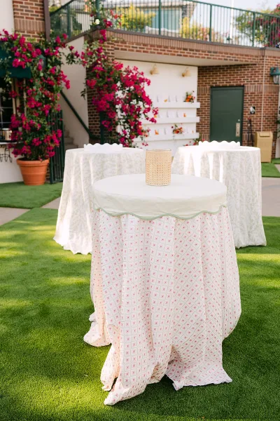 Cocktail table decor with patterned linens and a candle holder beside potted flowering vines on a brick patio with railing and lawn