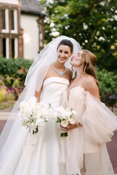Bride and bridesmaid share a kiss on the bride’s cheek, holding white rose and greenery bouquets, with a cathedral veil in a garden walkway.