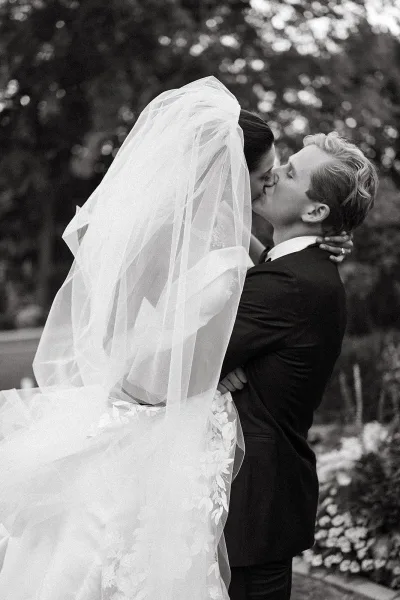 Wedding kiss in black and white as the bride and groom kiss under her long veil, lace gown details and garden trees softly blurred behind