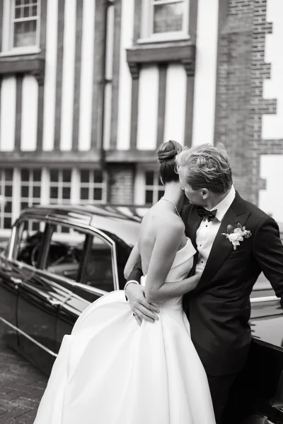 Wedding couple portrait in black and white as bride and groom hug with a forehead kiss beside a classic car on a street by a historic brick building facade