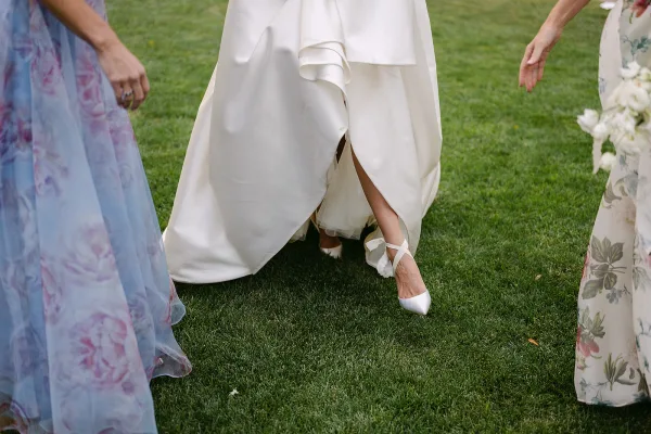 Wedding dress detail showing a satin bridal gown slit, white ankle-strap heels, and a tulle underskirt while walking on grass lawn