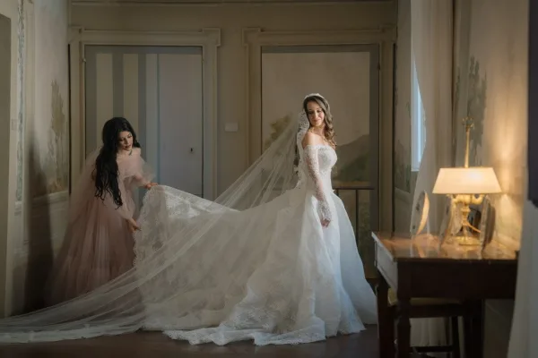 Bridal portrait of a bride in lace gown with a cathedral veil as a bridesmaid stands nearby before a painted wall mural and window.