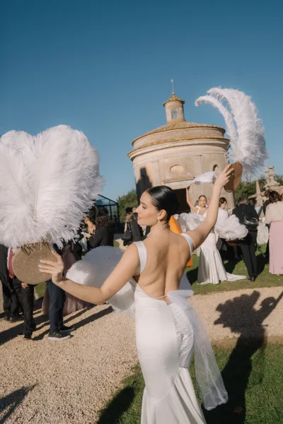 Wedding entertainment with feather fan dancers gliding on a lawn before a stone building, one wearing a pearl necklace beneath a blue sky.