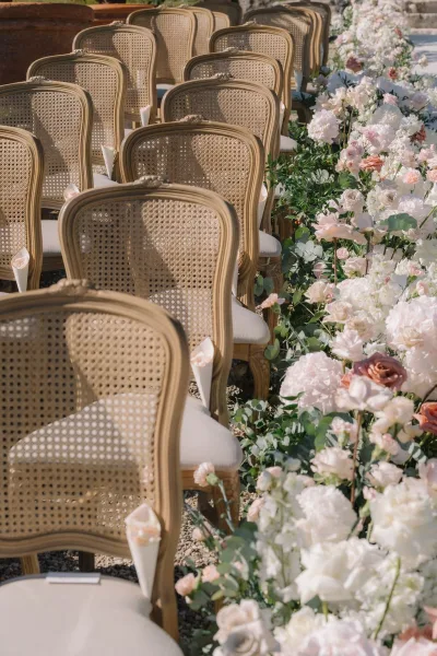 Ceremony aisle design featuring cane back wedding chairs flanking a neat aisle on gravel, with a grand stone planter and lush garden backdrop.