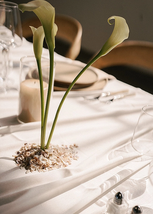 Reception tablescape with minimalist wedding tablescape styling, calla lilies, seashells, white linens, taper candles, and modern place settings in soft indoor light