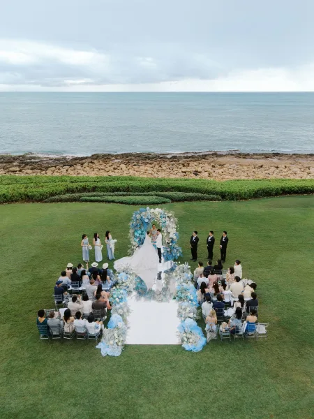 Ceremony moment with the couple at the altar on a mirrored aisle, an oceanfront lawn setting with rocky shore and cloudy sky beyond.