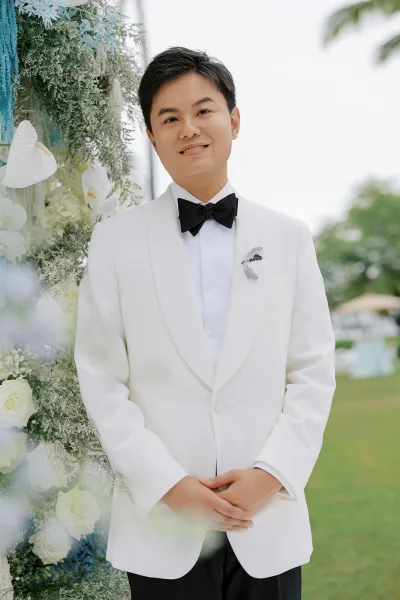 Groom portrait of a white tux groom smiling, a single boutonniere pinned to his jacket, standing on a garden lawn with trees under open sky.