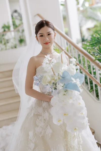 Bridal portrait of a bride with bouquet featuring cascading orchids, standing on a staircase with white walls and indoor plants.
