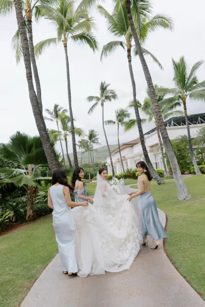 Bride with bridesmaids, the bridal party walking along a palm-lined garden path as she holds a white bouquet, mountains in the distance.