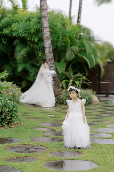 Flower girl portrait of a toddler flower girl wearing a flower crown, set amid tropical greenery and palm trees along a stone path.