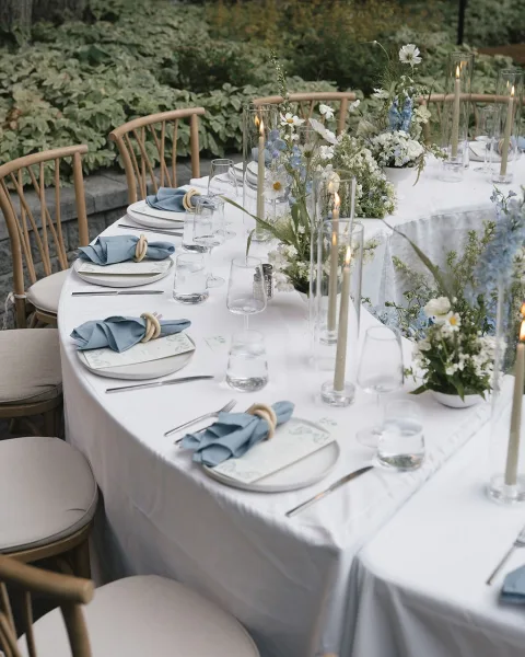 Reception tablescape with an outdoor reception table set in blue napkins, candles and floral centerpieces against garden greenery and stone ledge