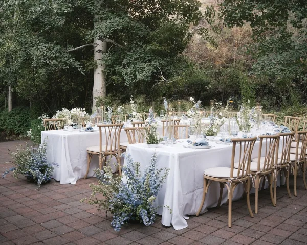 Reception tablescape with outdoor reception tables dressed in white linens, blue florals, taper candles, and glassware on a brick patio under trees