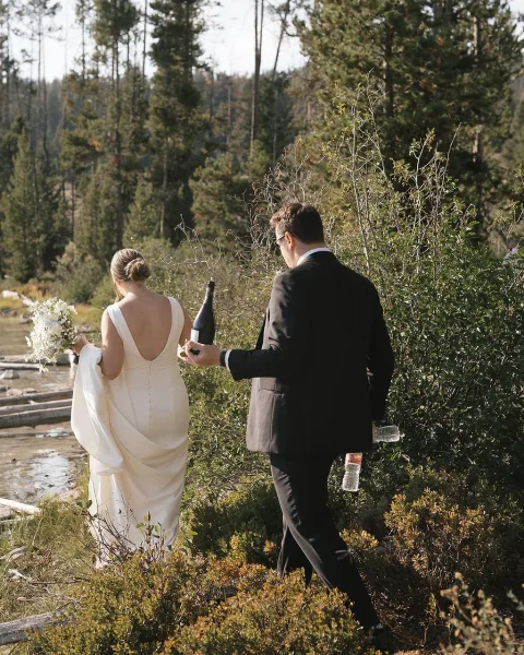 Couple portrait of bride and groom walking away along a rocky lakeshore, bride holding bouquet in a forest setting