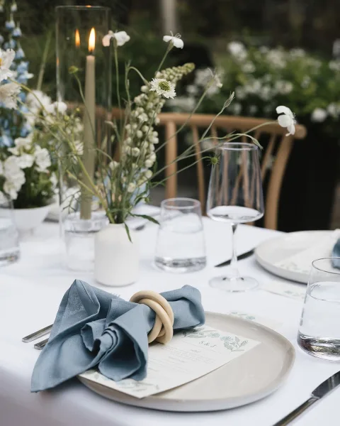 Reception tablescape with wedding place setting, linen napkin and taper candle, white floral centerpiece in greenery with wooden chairs