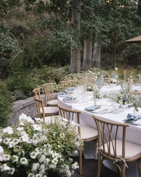 Outdoor reception tablescape with garden wedding reception table styling, blue napkins, taper candles, and floral centerpieces on a patio by trees